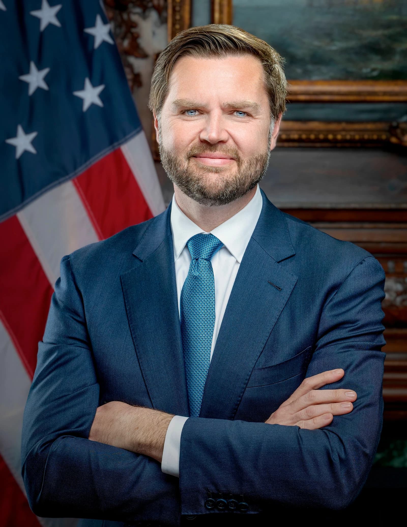 JD Vance, US Vice President, standing with arms crossed in front of an American flag.