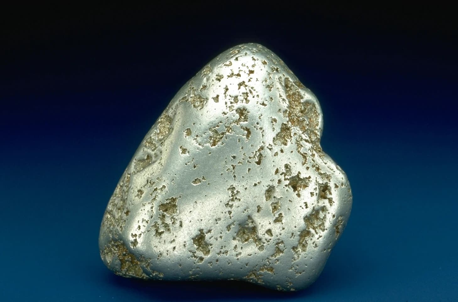 Close-up of a raw, irregularly shaped platinum nugget on a dark surface.