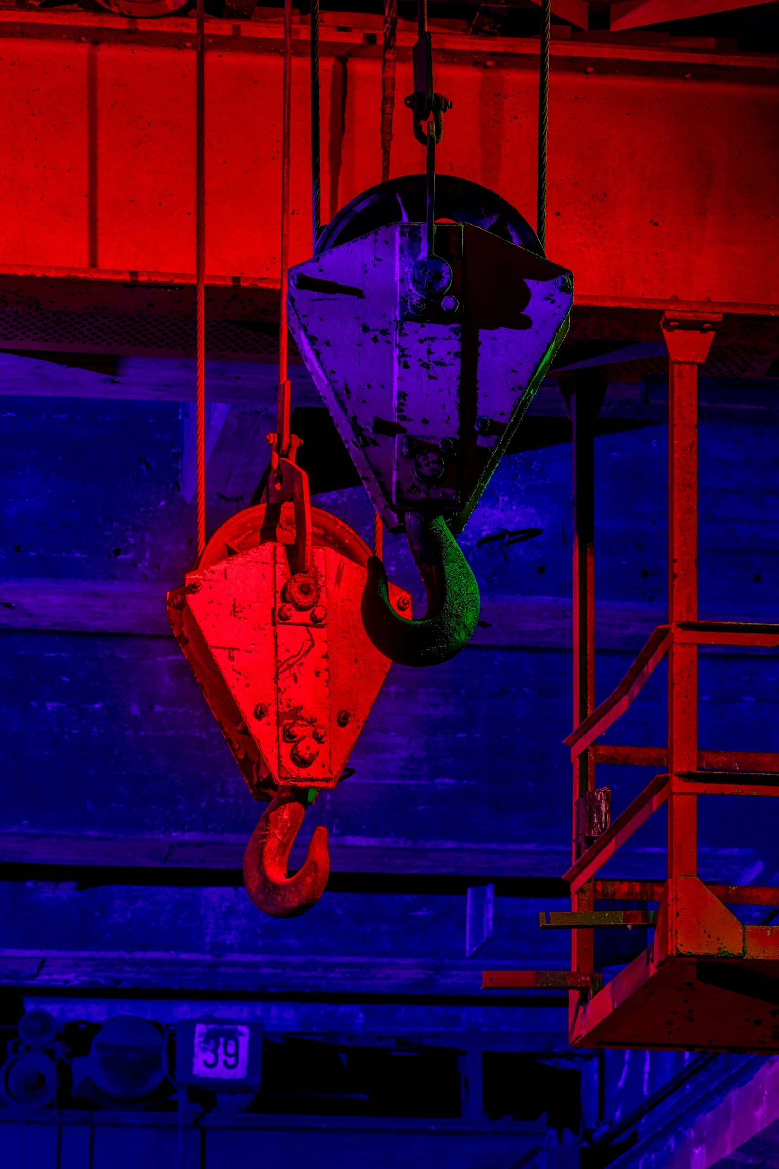 Close-up of two large industrial hooks hanging from machinery, illuminated by red and purple light.