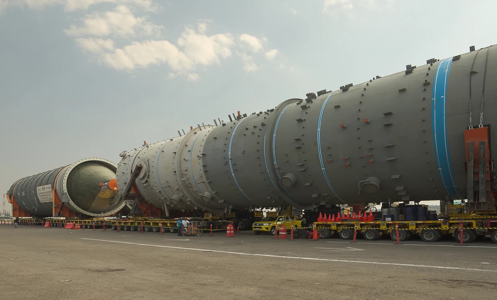 Massive gray distillation towers transported on multi-wheeled vehicles under a cloudy sky.