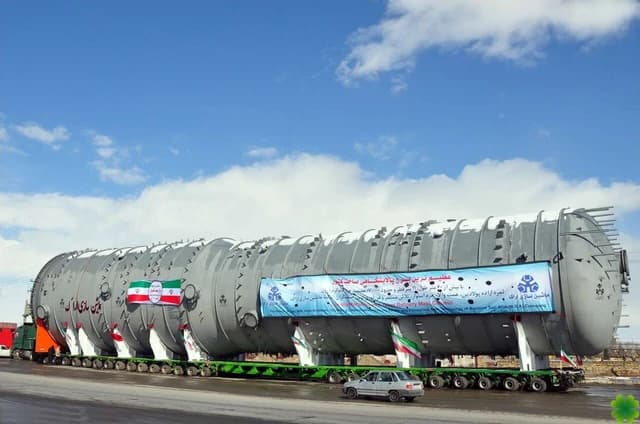 A giant distillation tower, a component of a refinery, being transported on a large truck.