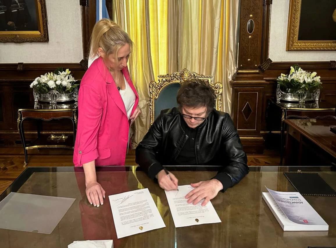 Javier Milei, President of Argentina, signs a document at a wooden desk.