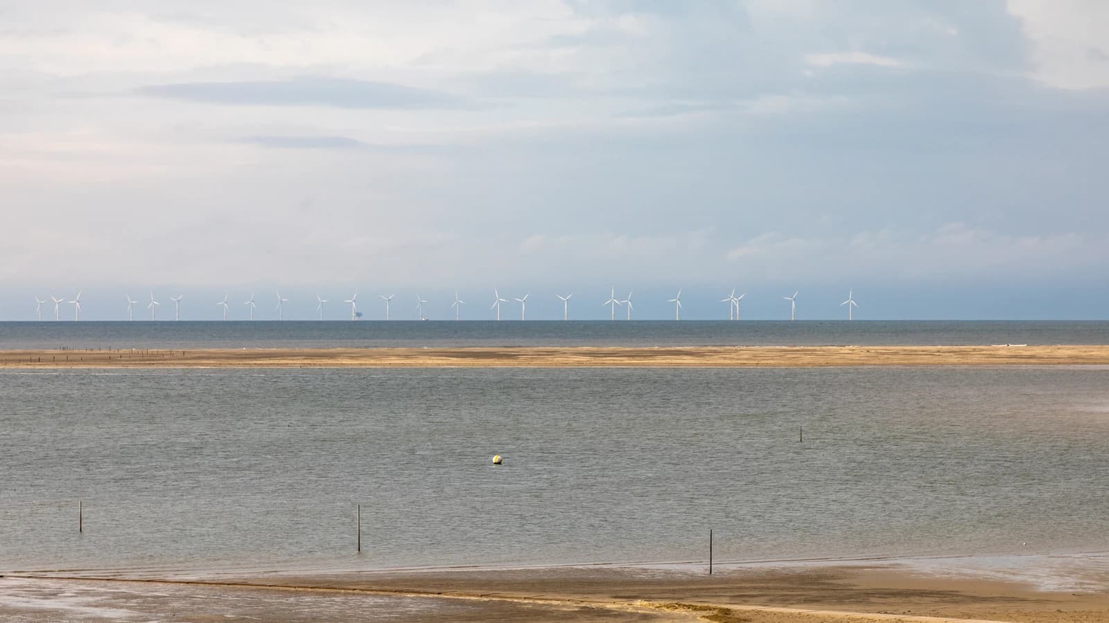 Offshore wind turbines line the horizon over the sea, viewed from a sandy beach.