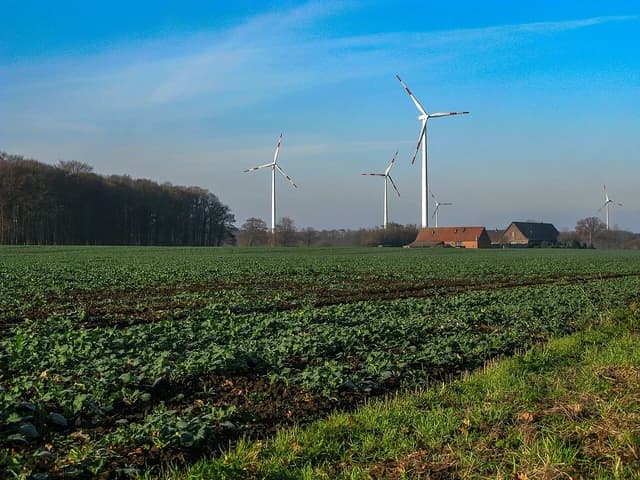 Several wind turbines stand tall in a green field with trees and buildings in the distance.