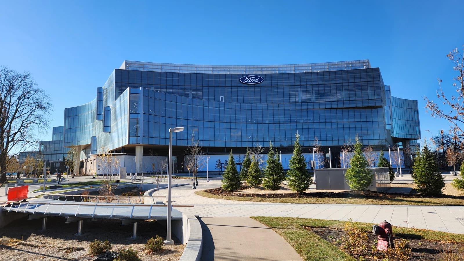 Ford World Headquarters building, a large glass structure, viewed from a sidewalk on a sunny day.