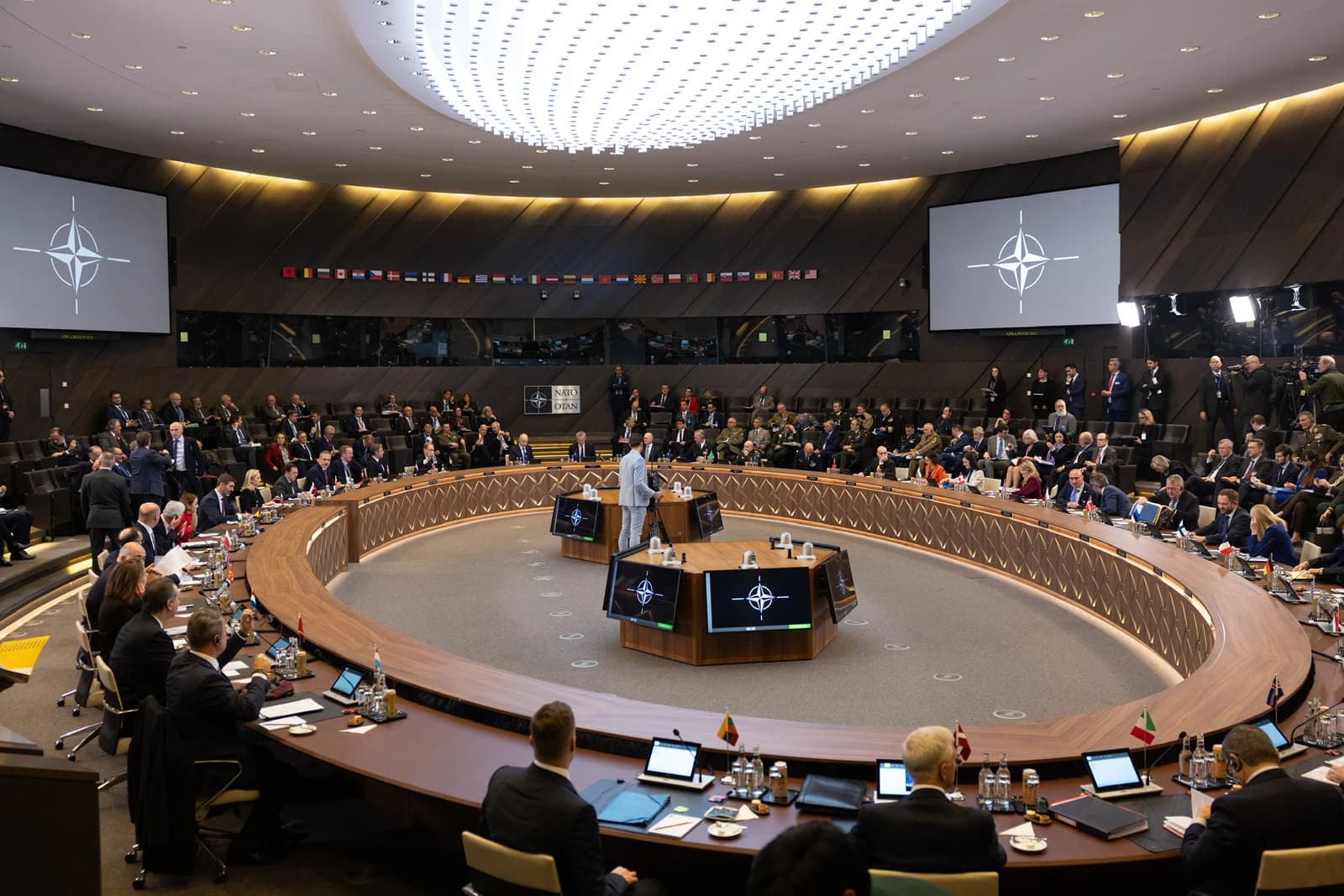 Secretary Blinken participating in a NATO Foreign Ministers Meeting, seated at a long, curved table.