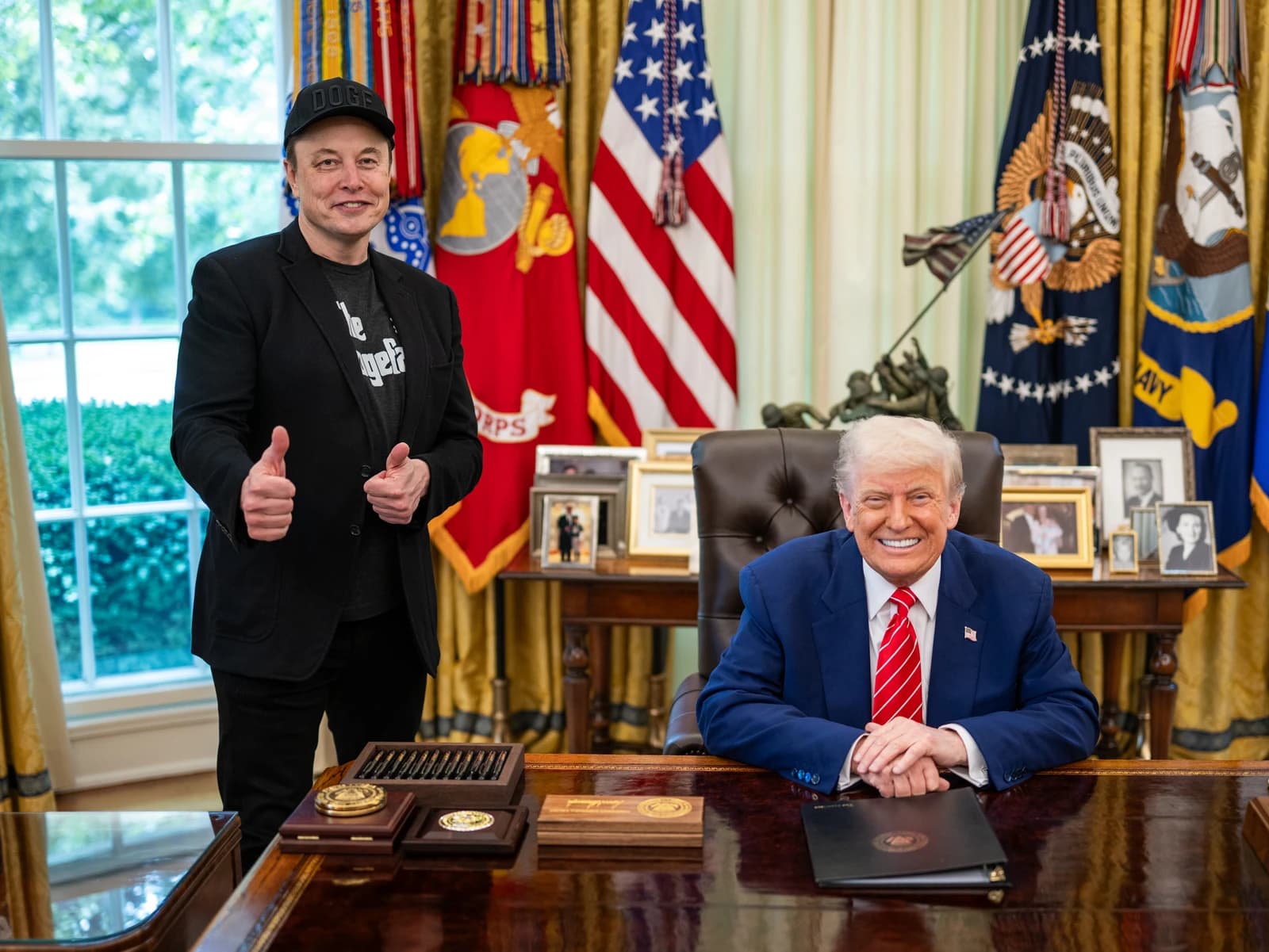 Donald Trump smiling behind a desk, Elon Musk standing next to him with thumbs up.