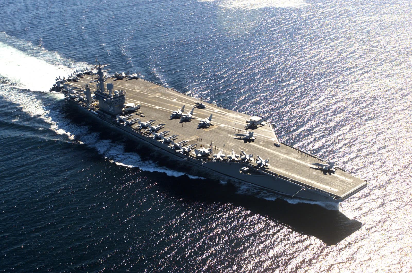 An aerial view of the USS Nimitz aircraft carrier with several jets on its deck, moving through water.