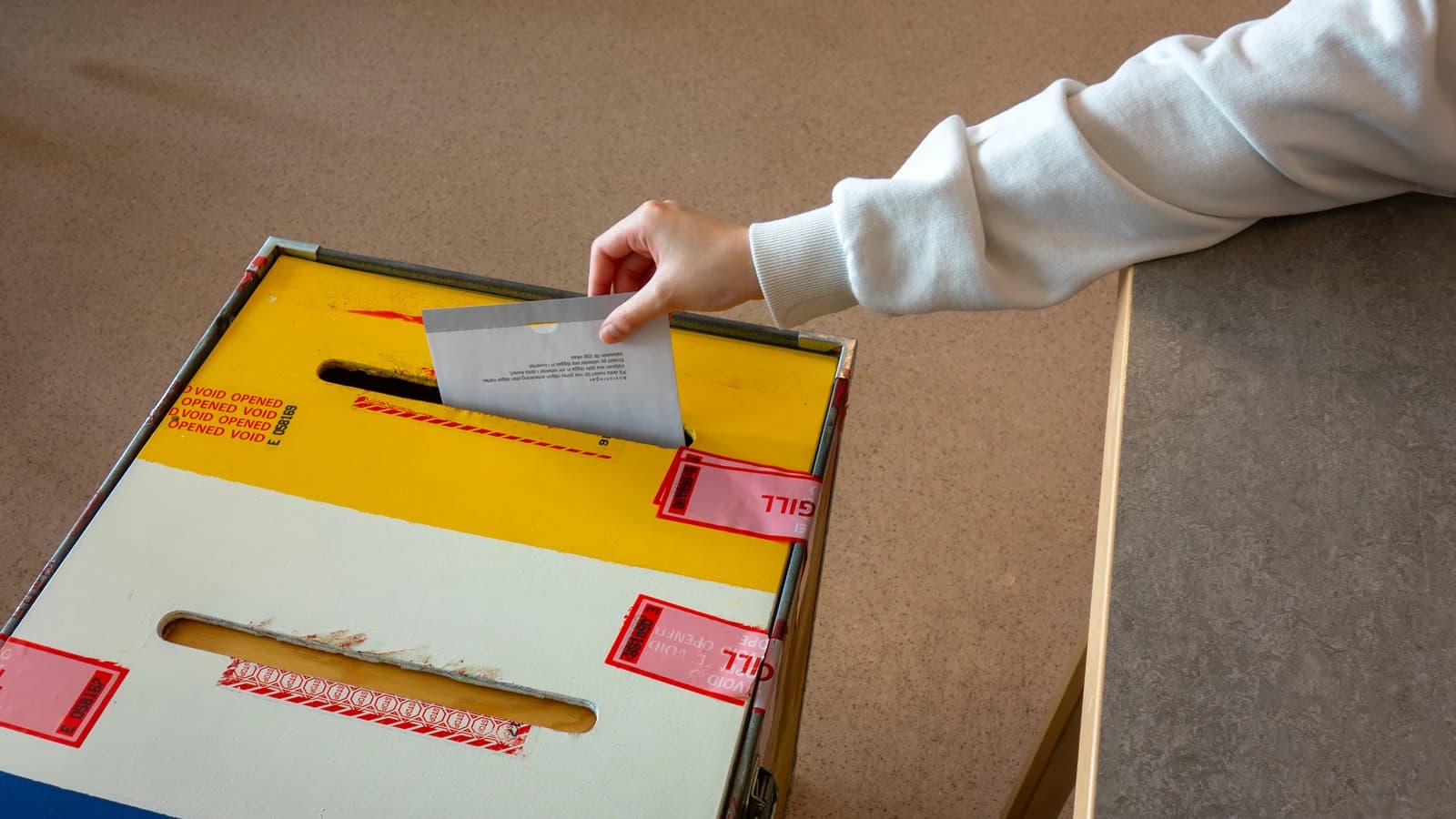 A person's hand deposits a ballot into a yellow and white ballot box.