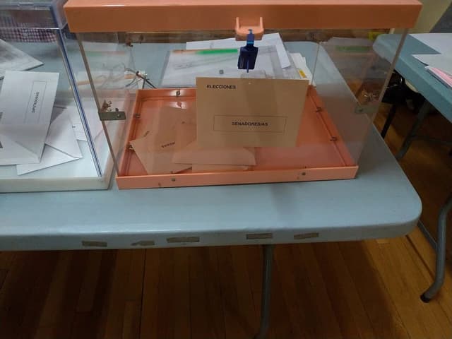 Transparent ballot box with 'SENADORES/AS' sign on a table.