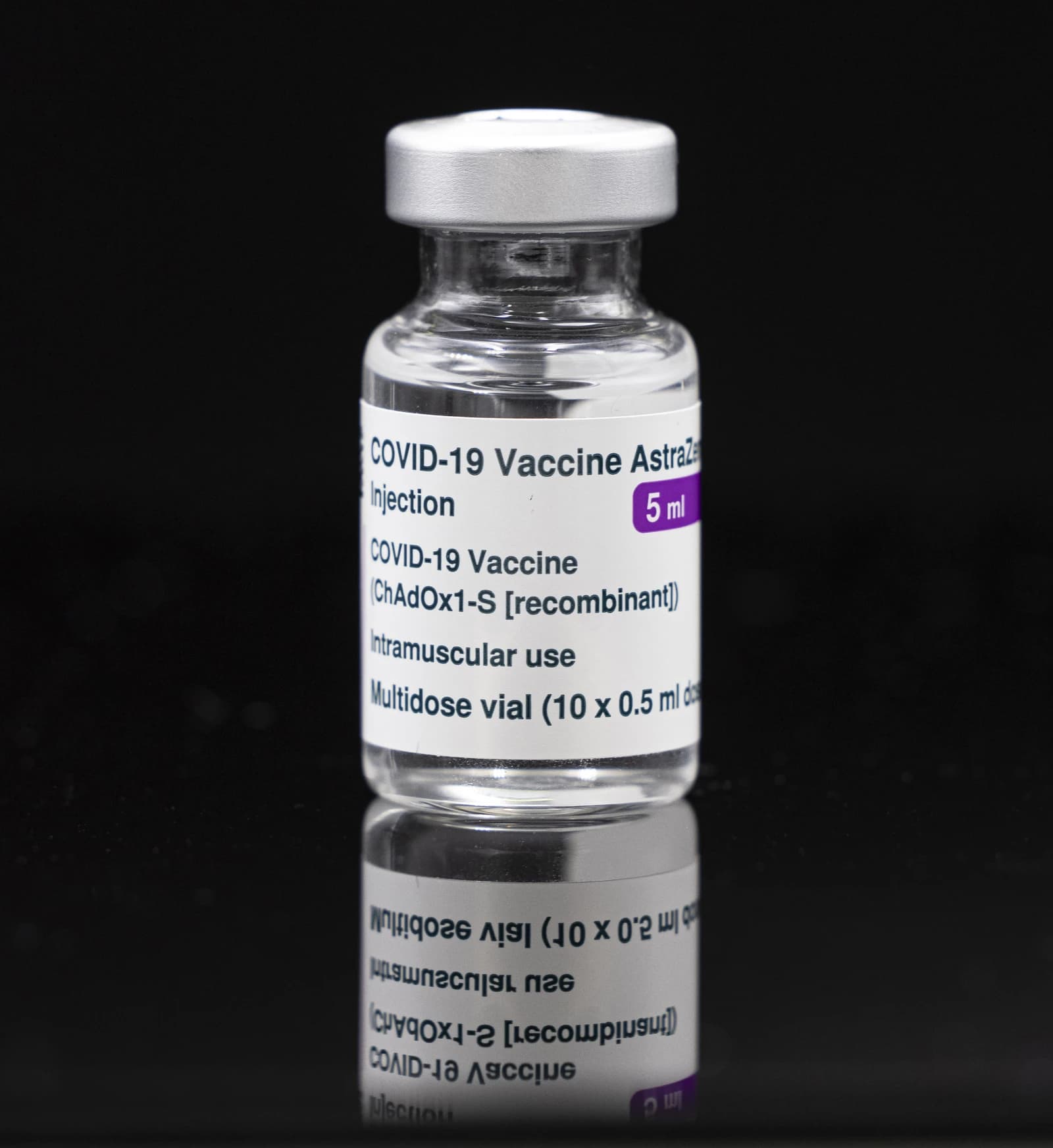 A clear glass vial labeled 'COVID-19 Vaccine AstraZeneca injection' stands on a reflective surface.