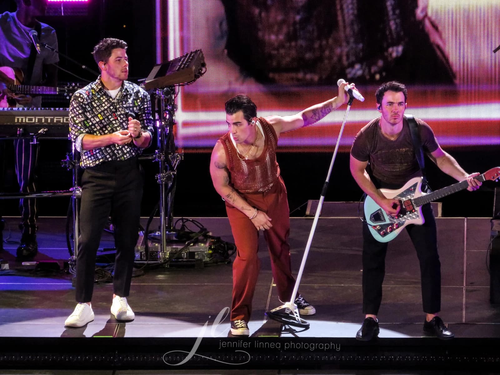 Kevin, Joe, and Nick Jonas performing on stage with microphones and a guitar.