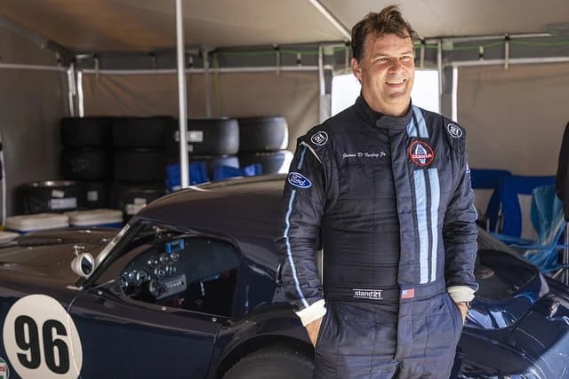 Jim Farley, CEO of Ford, standing in a race suit next to a Shelby Cobra race car.