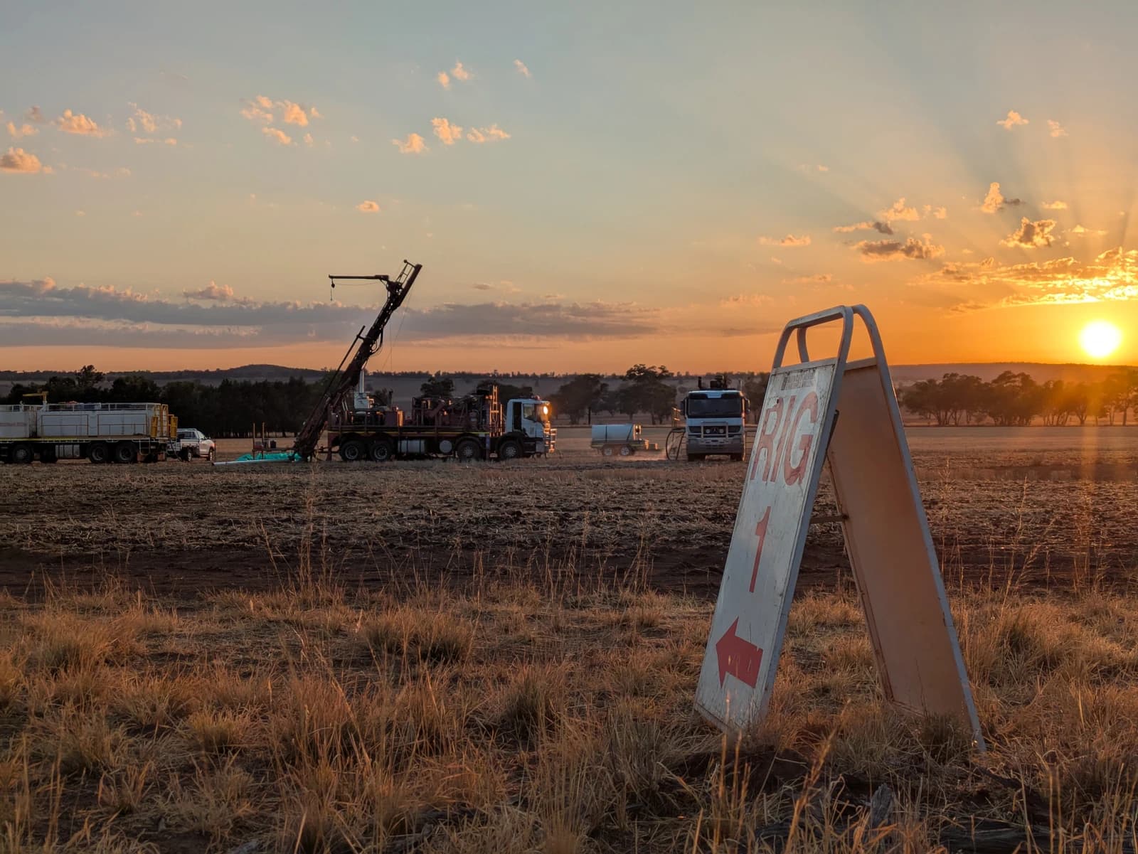 Drilling rig and trucks at a mining operation under a sunset sky.