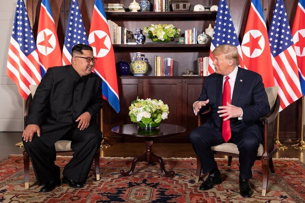 Donald Trump and Kim Jong-un sitting opposite each other, talking, with US and North Korean flags behind them.