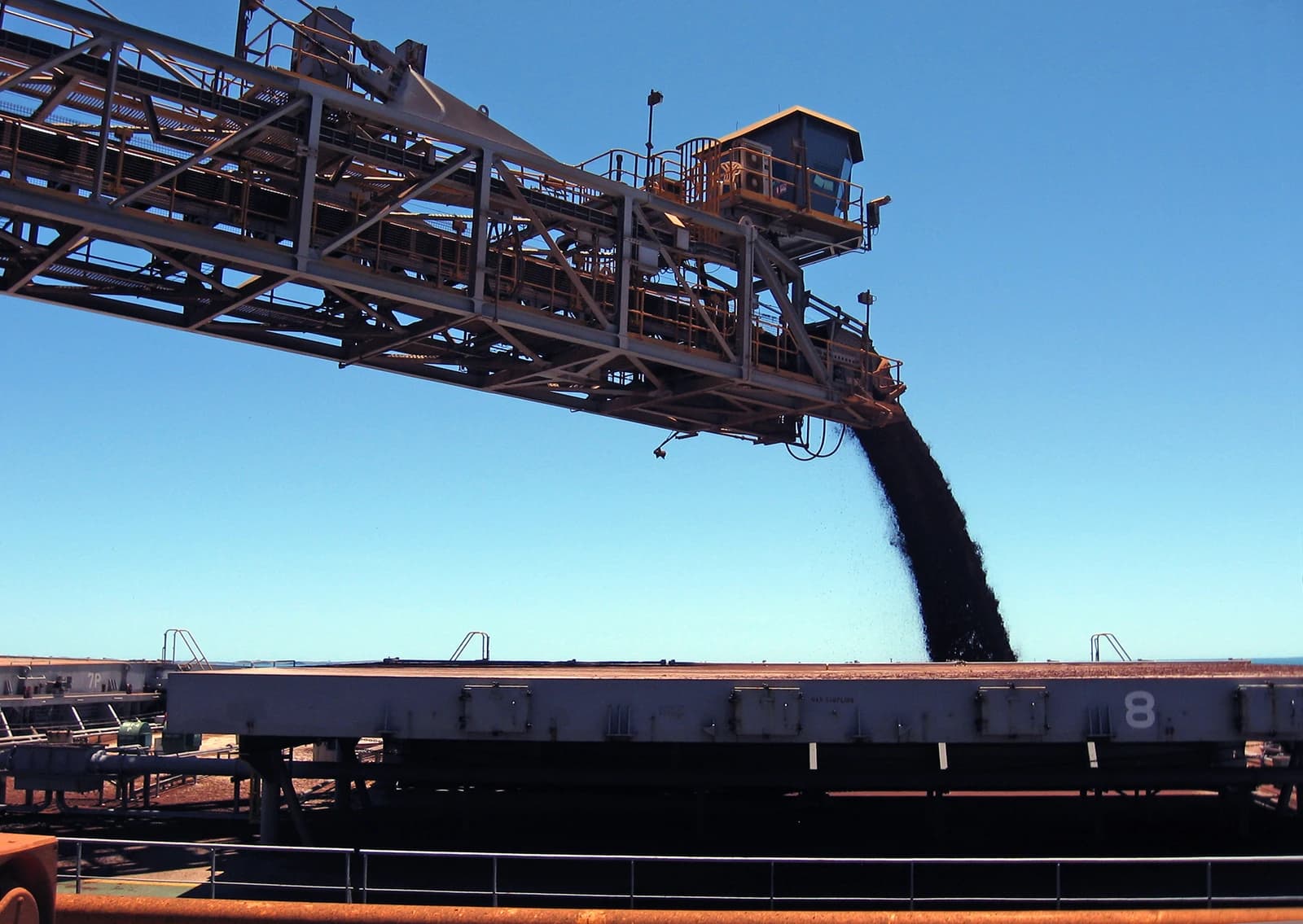 A shiploader discharges iron ore onto a large industrial platform under a clear blue sky.