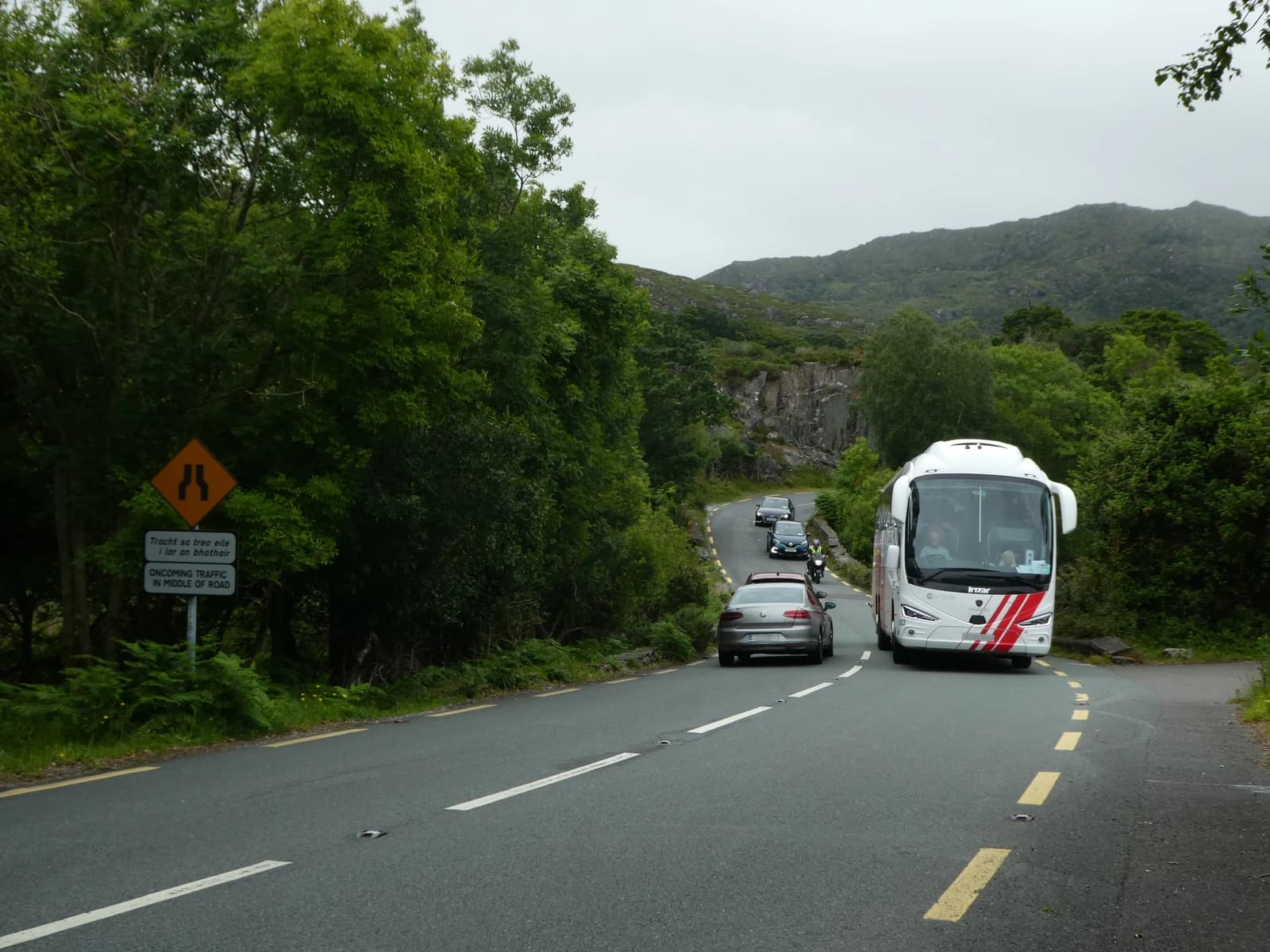 A tour bus drives on a narrow, winding road in a green, mountainous landscape in Ireland.