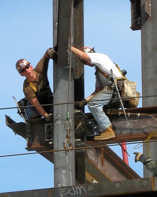 Construction workers on steel beams, one looking towards the camera, another working on the structure.