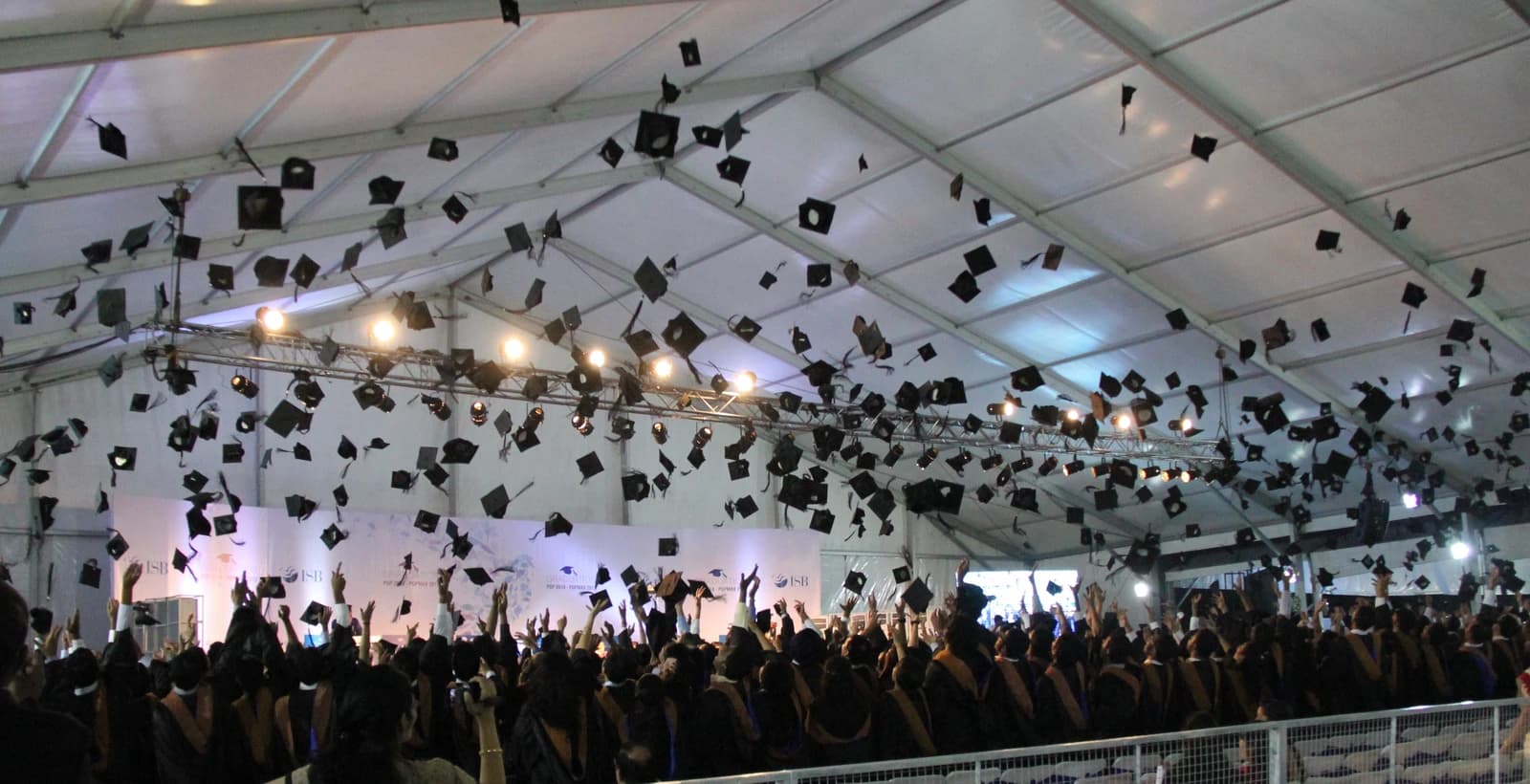 Graduates in academic regalia toss square academic caps into the air indoors.