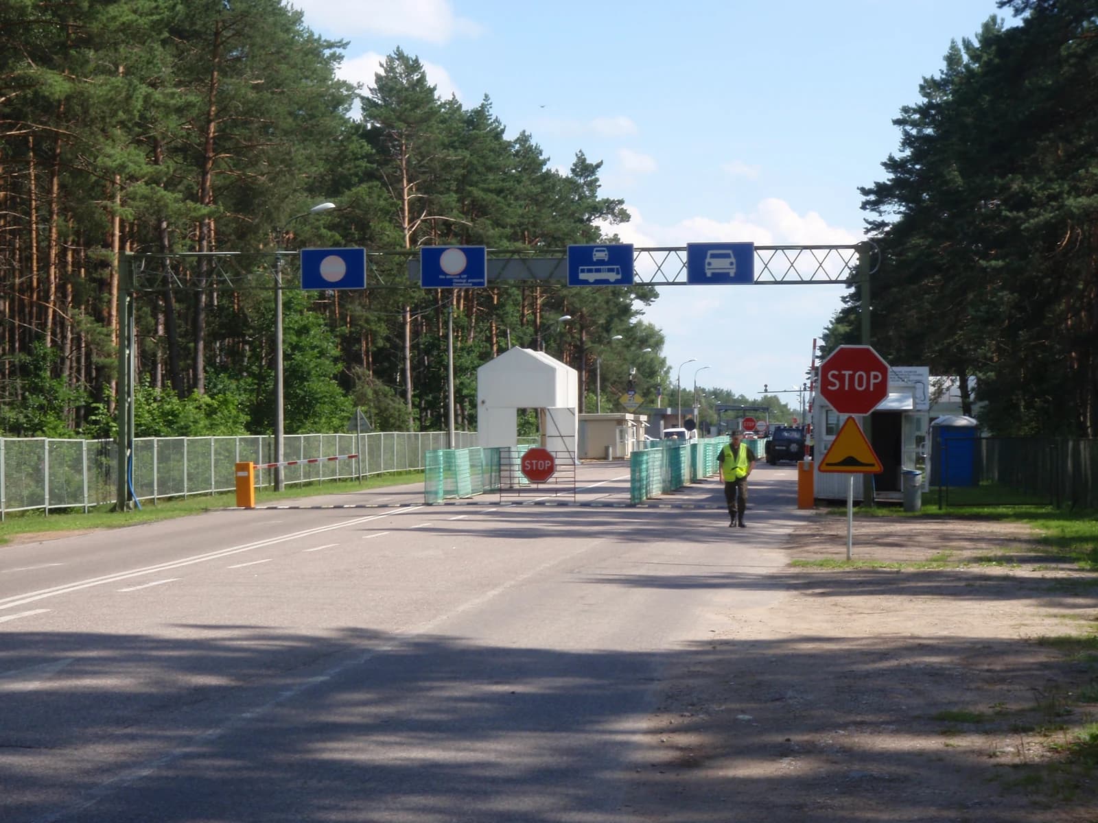 A border checkpoint with a uniformed person, stop signs, and barriers on a road.