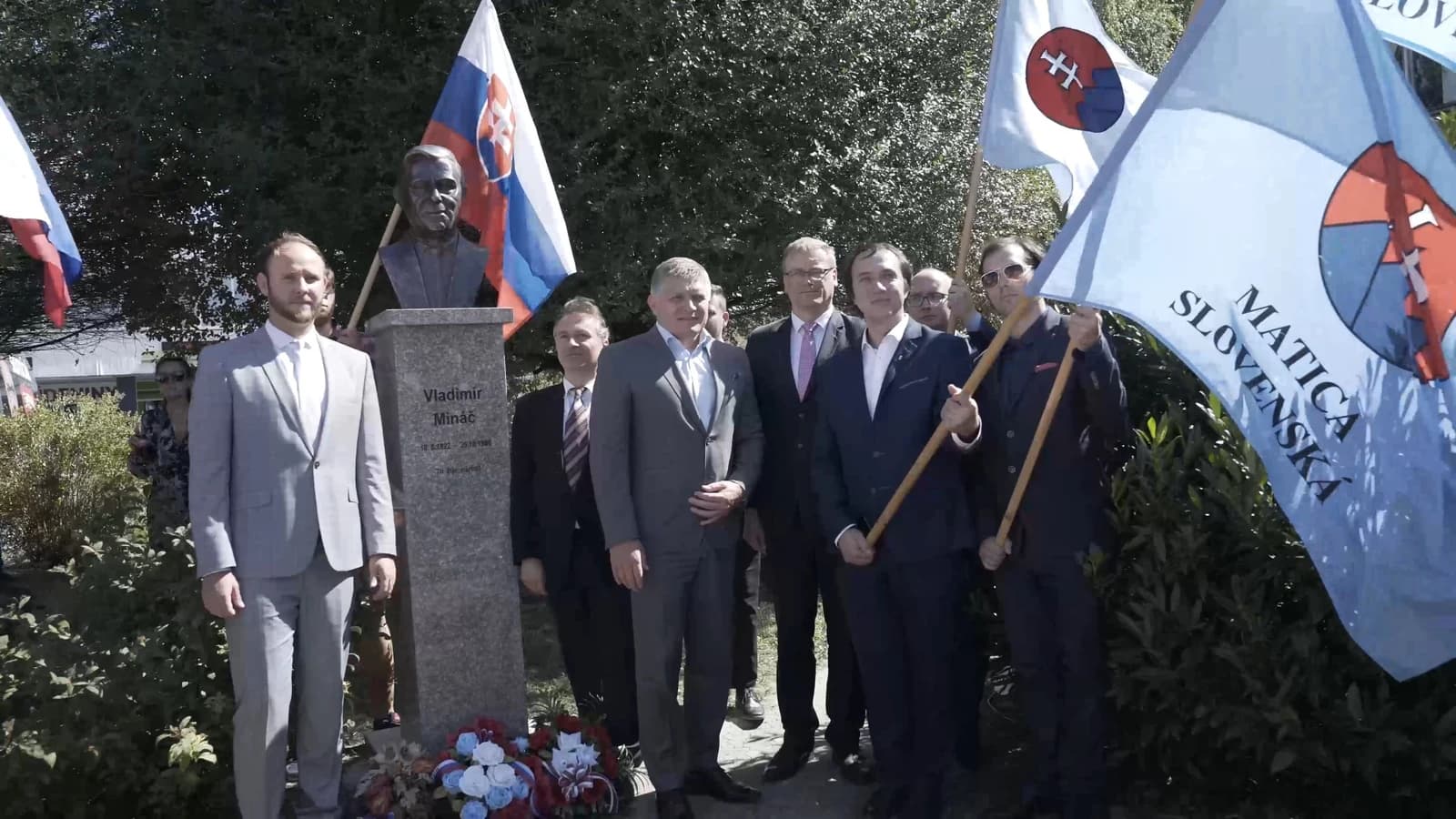 Robert Fico, a Slovak politician, stands with other men near a bust and flags.
