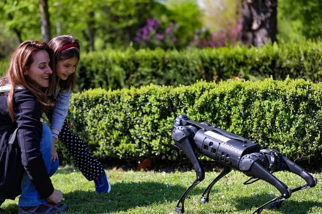 Woman and child kneeling and looking at a black Unitree robot dog on a grassy lawn.