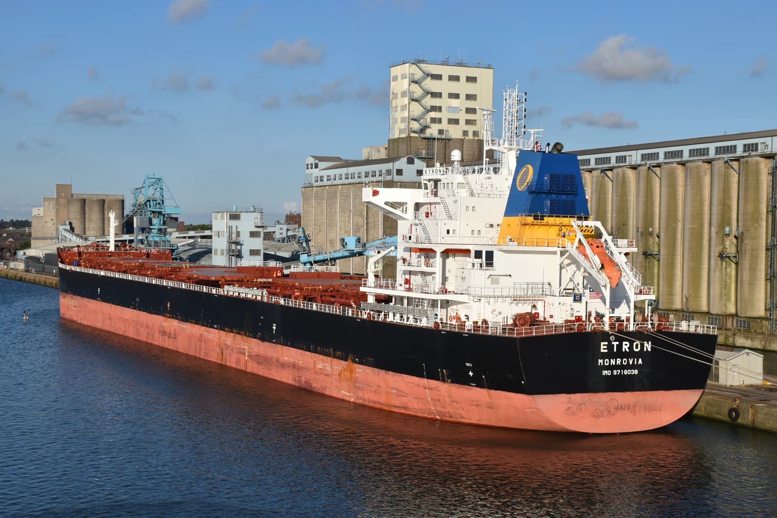 The cargo ship Etron docked in a port with large industrial silos behind it.