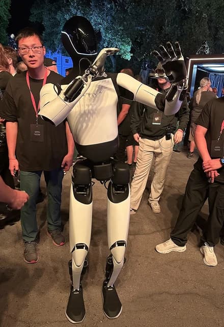 Tesla Optimus humanoid robot stands with arm raised among attendees at an event.
