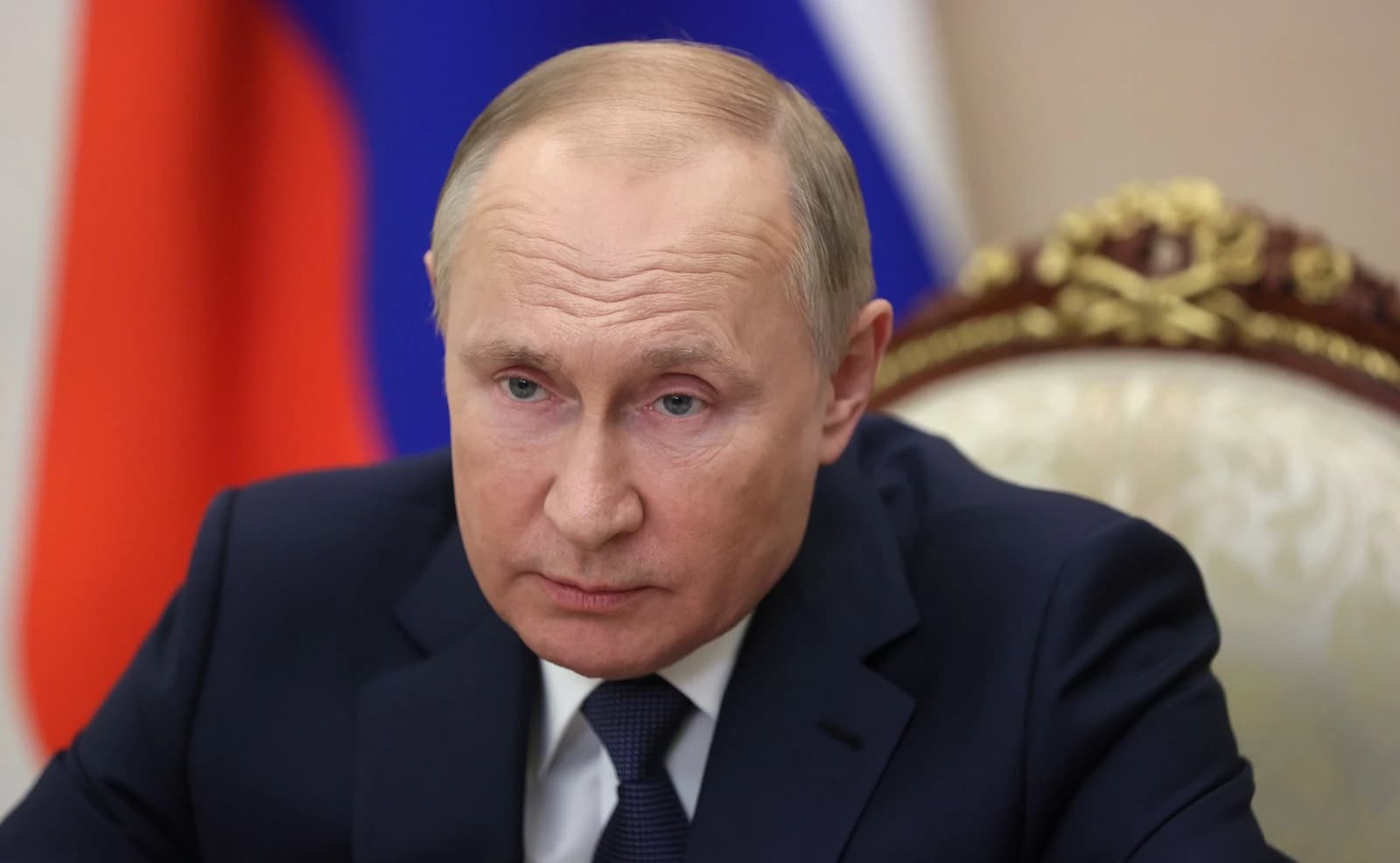 Vladimir Putin, President of Russia, sits in an ornate chair with the Russian flag behind him.