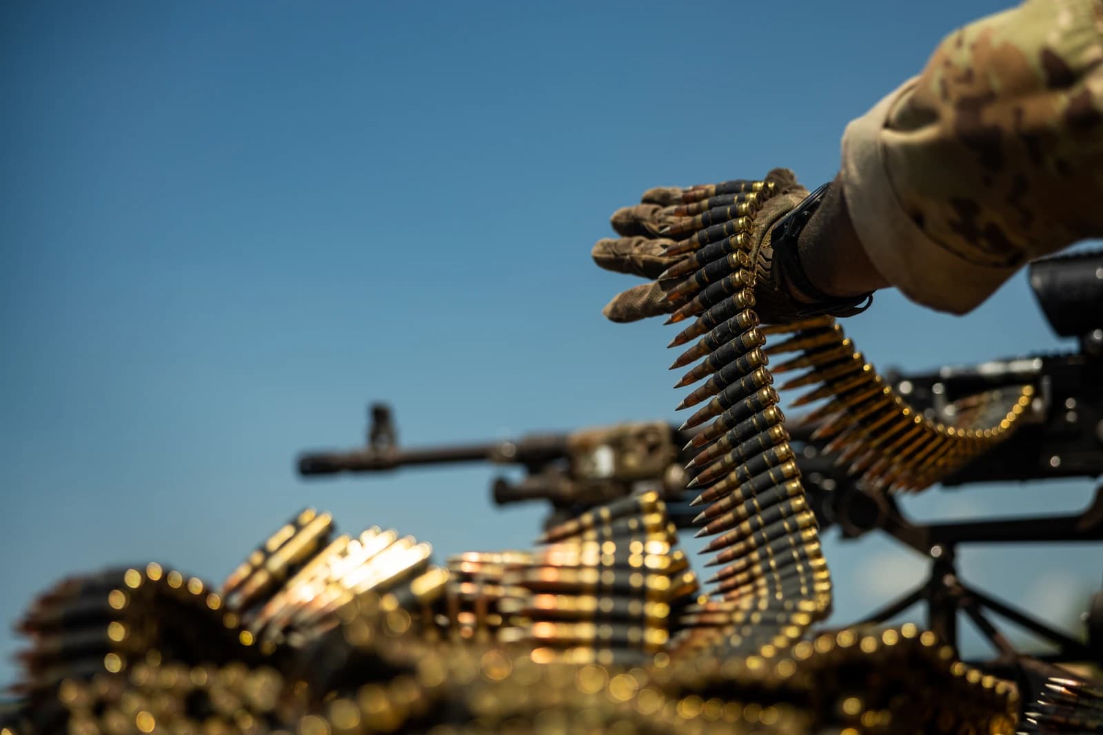 A gloved soldier's hand holds a long belt of ammunition, with a firearm in the background.