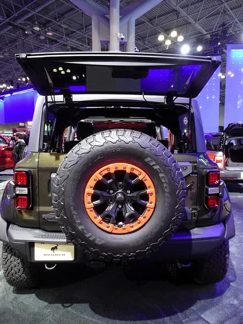 Rear of a green Ford Bronco Raptor with orange wheel accents and open tailgate.