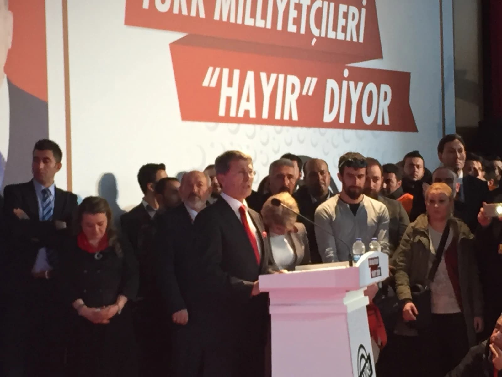 Man in a dark suit and red tie speaking at a white podium, surrounded by people.