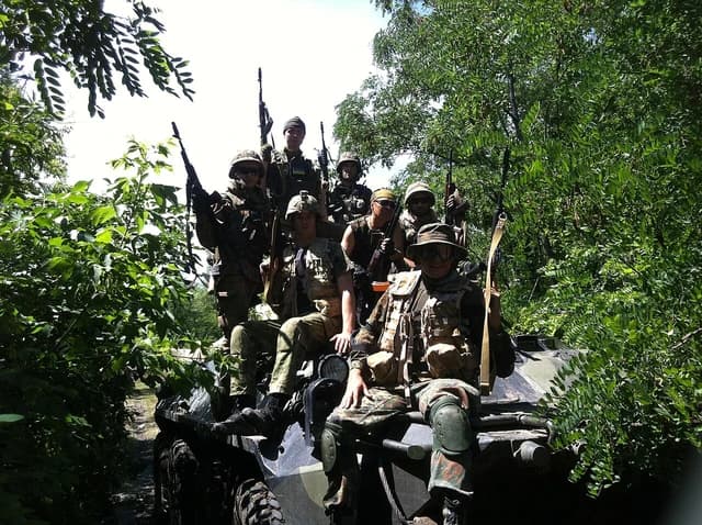 Soldiers wearing camouflage and helmets pose on an armored vehicle surrounded by foliage.