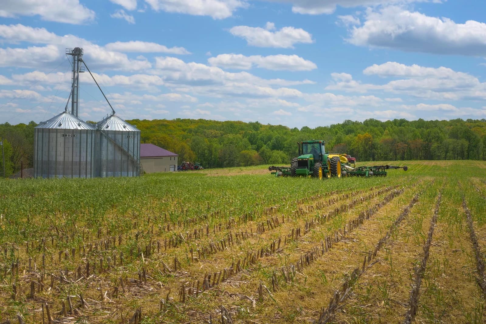 Farm field with grain storage silos and a green tractor in the distance.