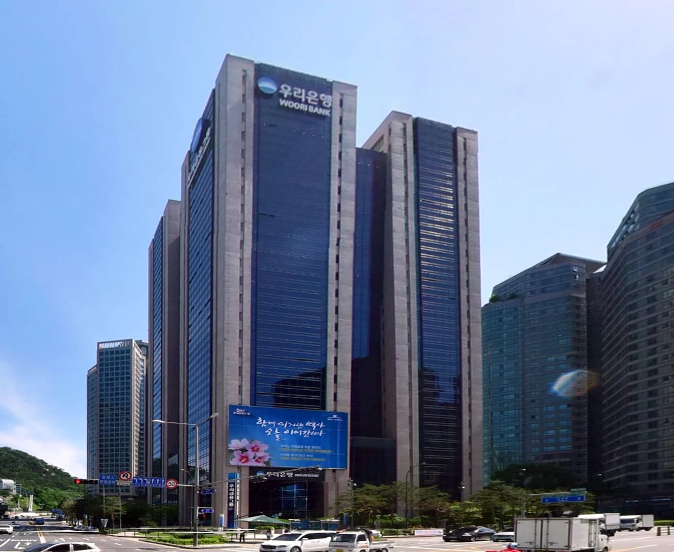 Woori Bank headquarters building stands tall among other buildings with busy street traffic below.