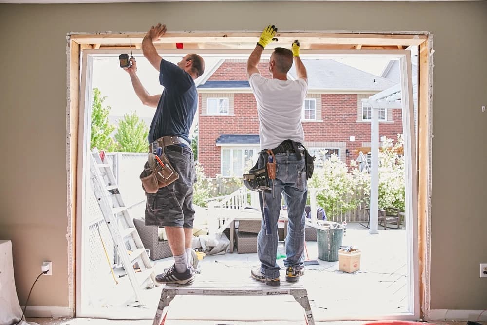 Two construction workers install a large window frame in a residential building.