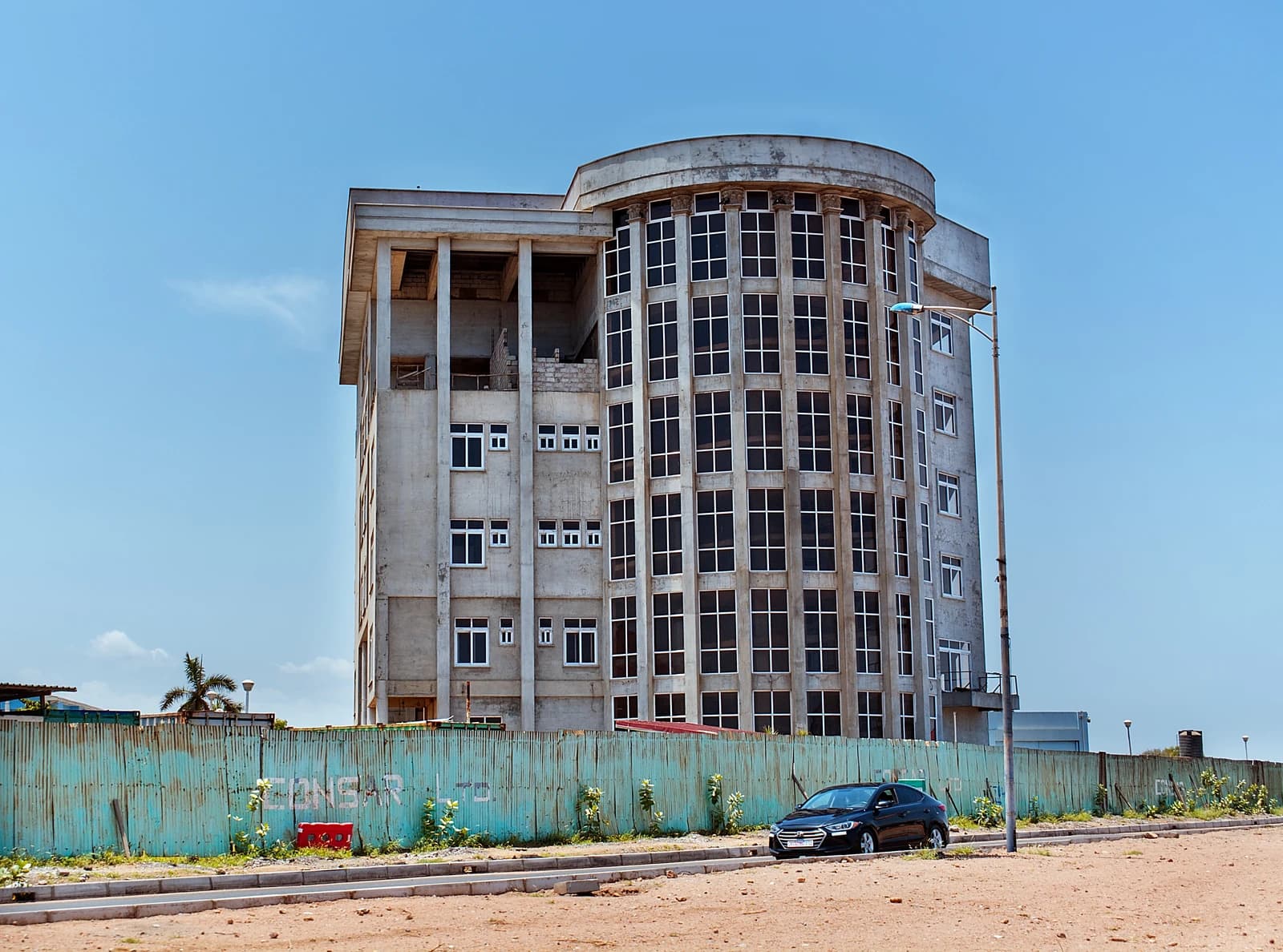 A modern building under construction, partially behind a green fence, with a black car.