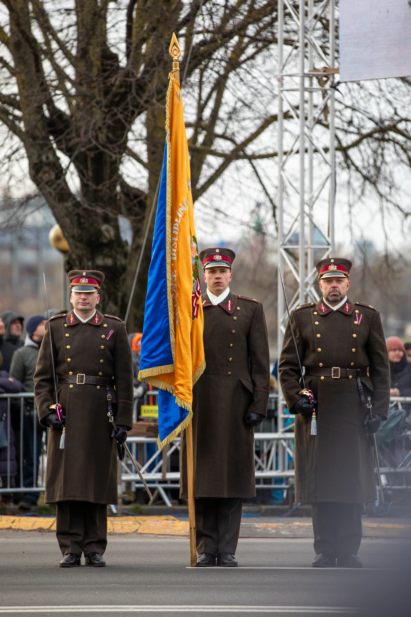 Three military officers in dark uniforms stand with a flag at a parade in Latvia.