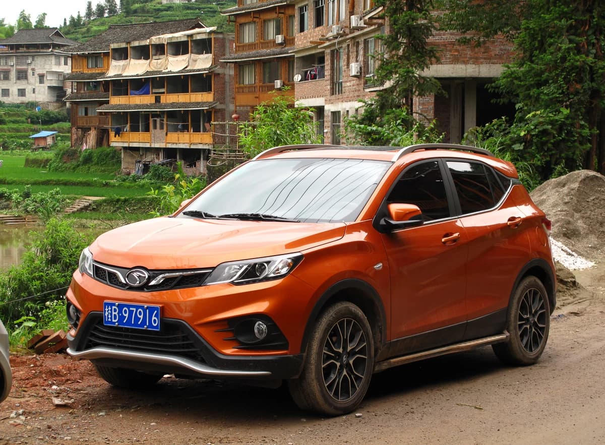 Orange Soueast DX3 compact SUV parked on a dirt road by traditional buildings.