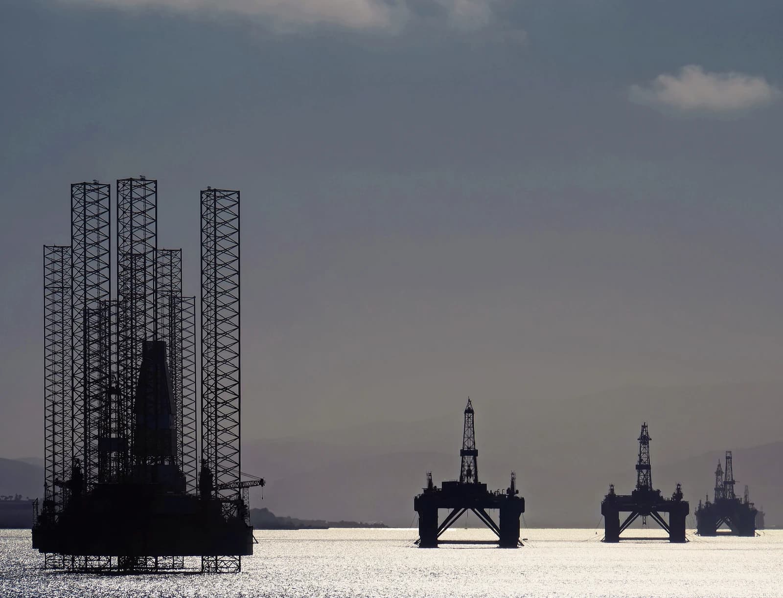 Multiple oil rigs anchored in a body of water, silhouetted against a bright sky.