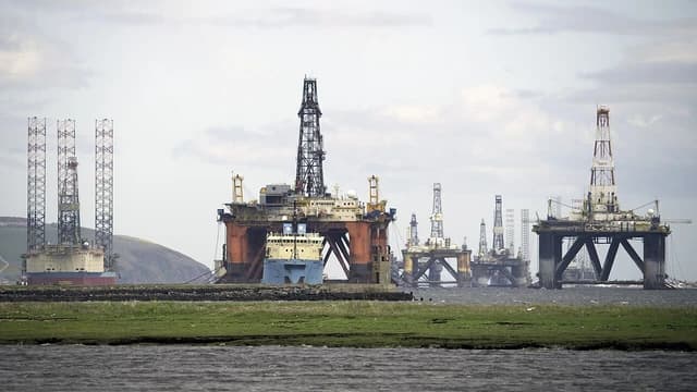 Several offshore oil rigs, large and complex structures, are visible from a grassy shoreline.