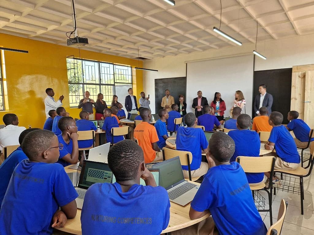 Students in blue shirts with laptops looking towards adults speaking in a smart classroom.