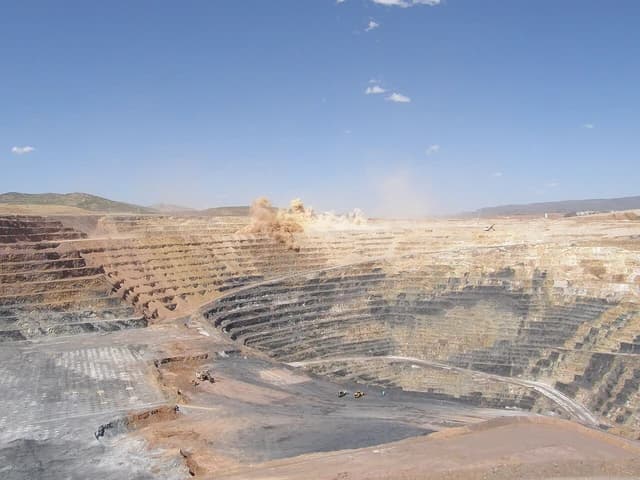 An open-pit mine with a controlled blasting operation creating a cloud of dust.