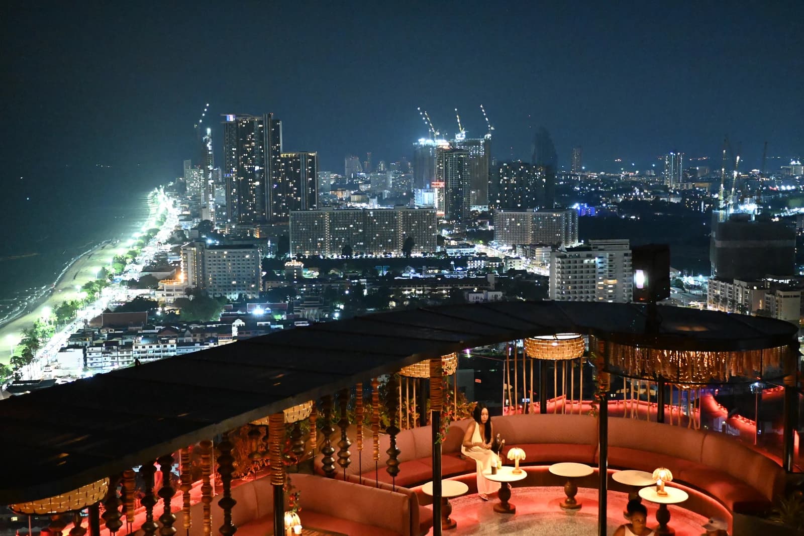 Woman relaxing in a rooftop lounge overlooking the illuminated Pattaya coastline at night.