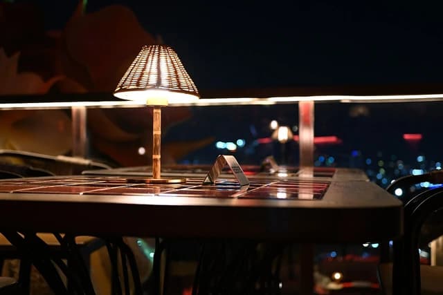 A lit table lamp on a rooftop dining table overlooking city lights at night.