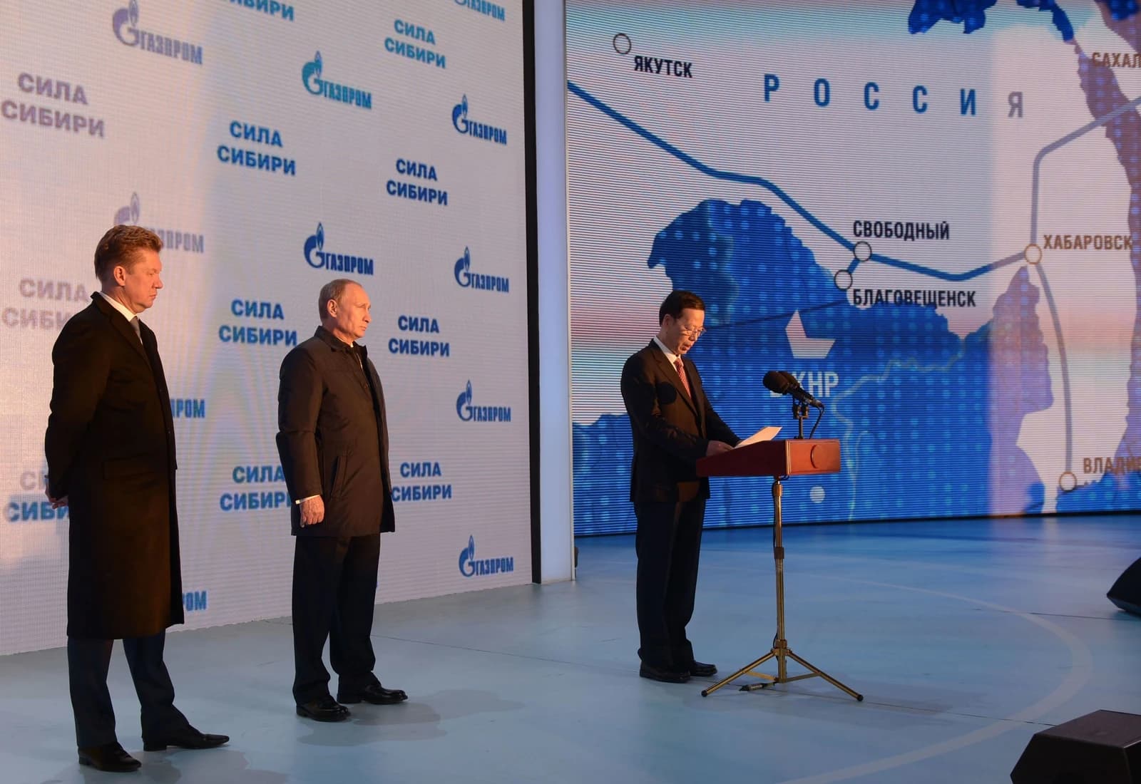 Vladimir Putin and Alexey Miller standing on stage, a map of the Power of Siberia pipeline behind them.