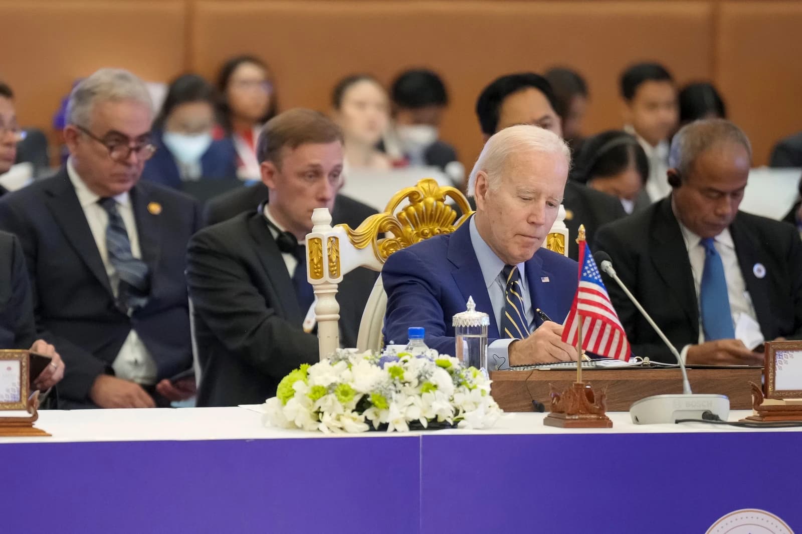 President Joe Biden sits at a long table, writing notes at an international summit.