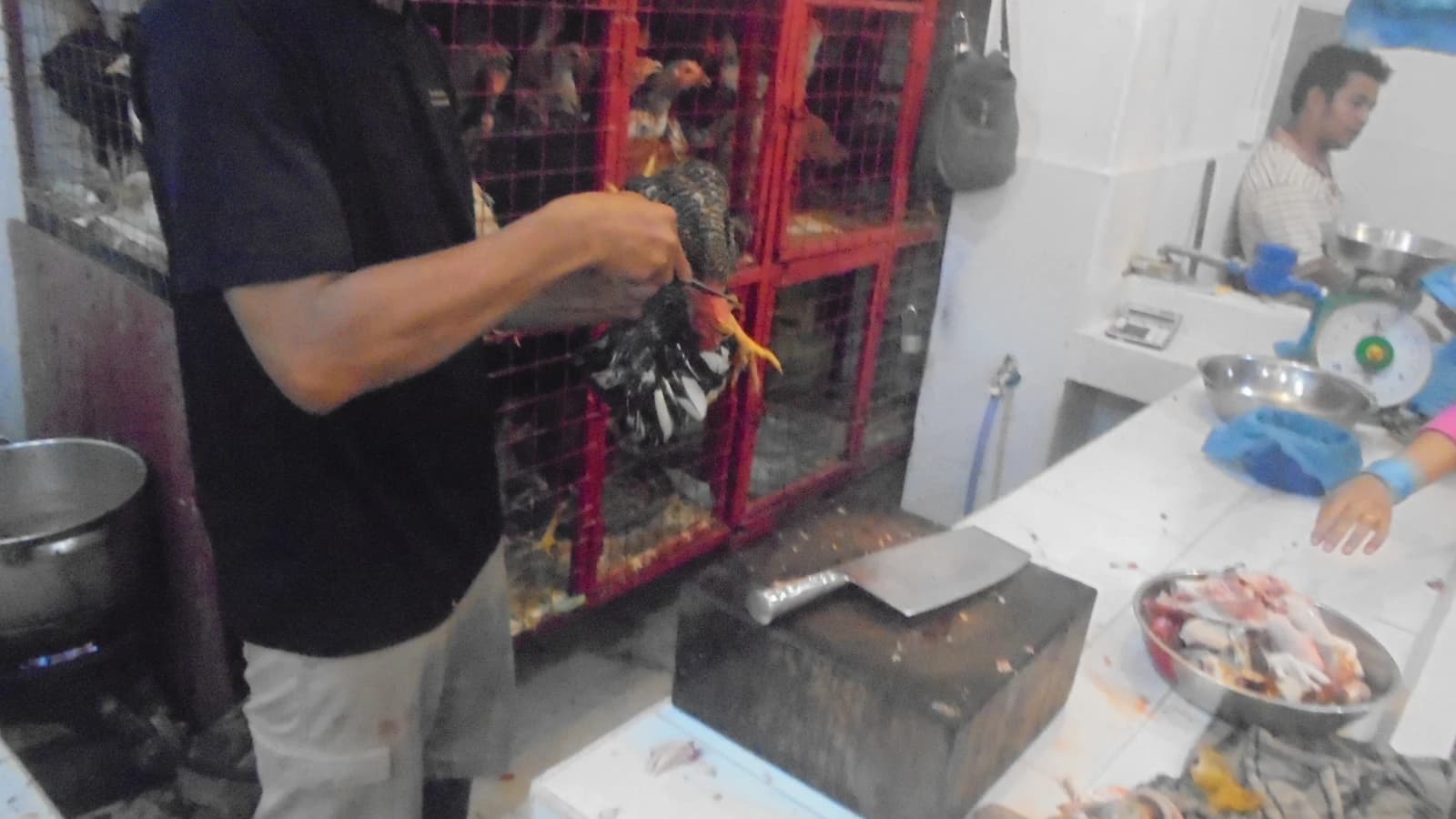Market vendor holding a chicken above a cutting board in a market stall.