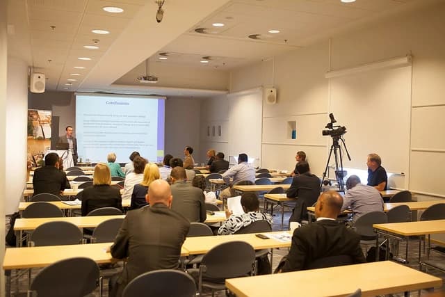 People attending a presentation in a conference room, facing a speaker and projected screen.