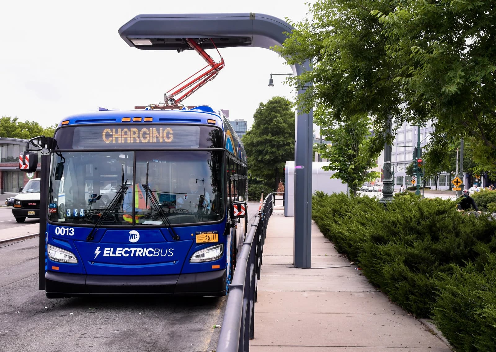 Blue electric bus connecting to an overhead charging station on a city street.